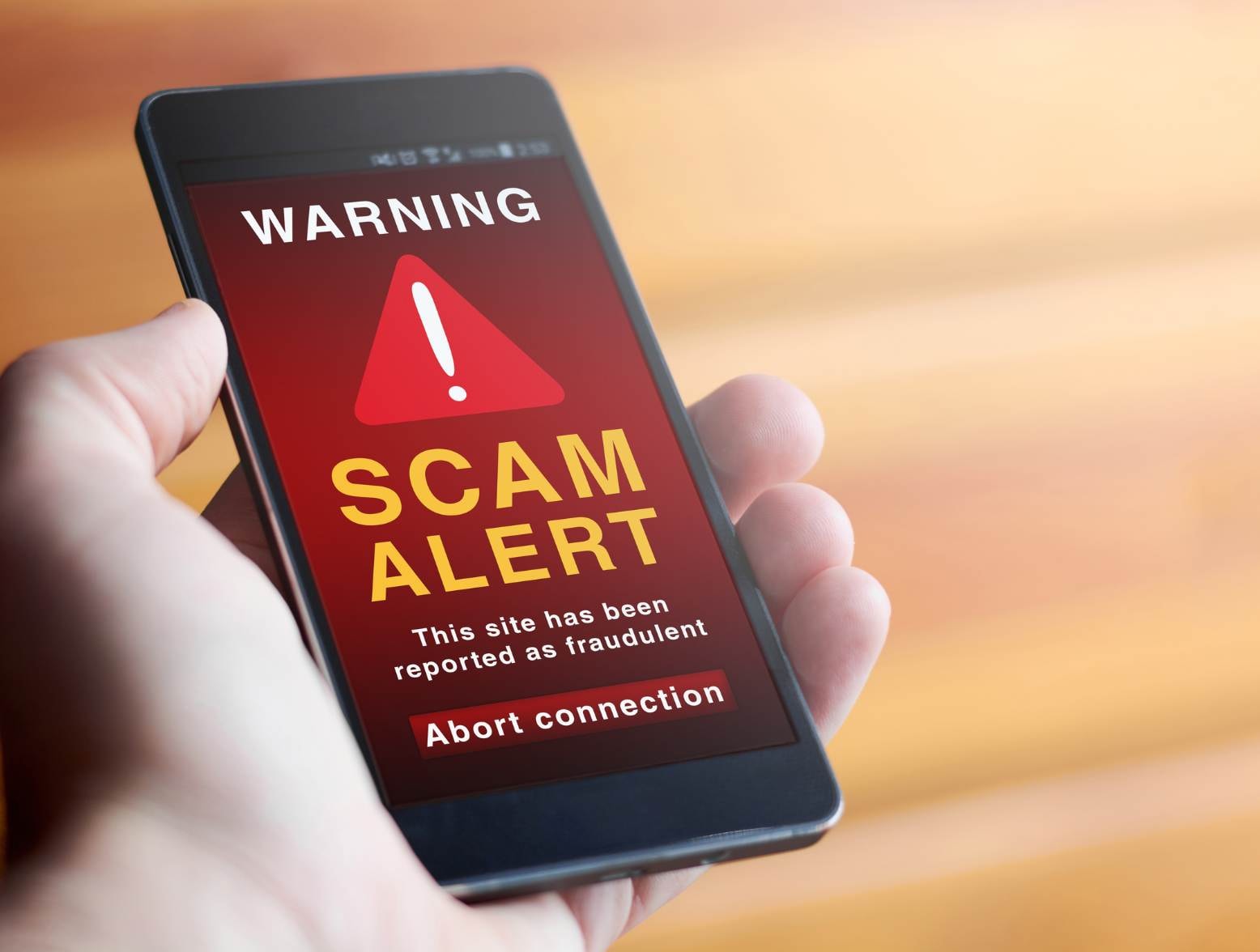 Closeup of male hand holding modern mobile phone with red screen and warning icon with the text SCAM ALERT This site has been reported as fraudulent.